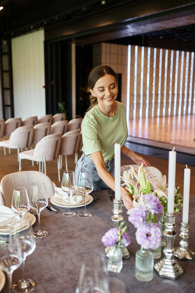 mladý dekoratér usmívá u stolu se slavnostním nastavením, květiny a svíčky v slavnostní síni - Fotografie, Obrázek