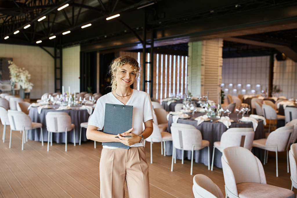 smiling blonde event manager with clipboard looking at camera in banquet hall with decorated tables - Photo, Image
