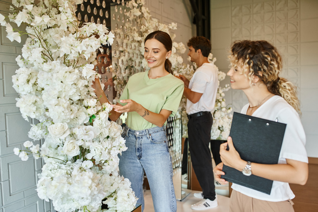 glücklicher Florist zeigt auf weiße florale nahe Teamleitung mit Klemmbrett in der Veranstaltungshalle, kreative Arbeit - Foto, Bild