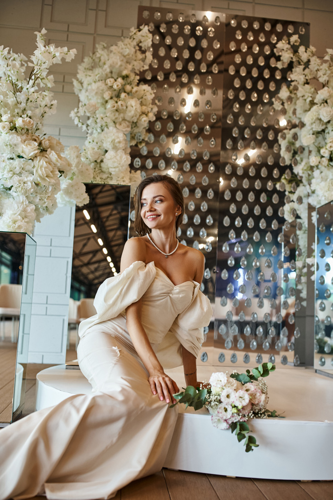 alegre joven novia en vestido de novia blanco sentado cerca de la decoración floral blanca en la sala de celebración - Foto, imagen