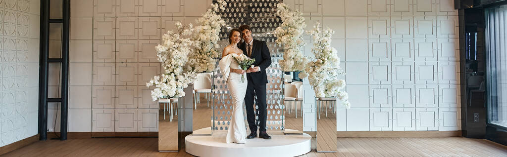 longueur totale des jeunes mariés élégants posant dans la salle d'événement décorée avec des fleurs blanches en fleurs, bannière - Photo, image