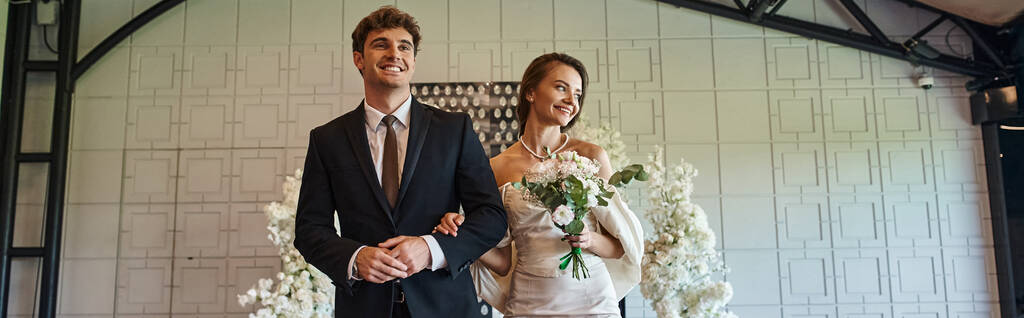 щаслива молода пара у весільному вбранні, що йде в сучасному залі подій з білим квітковим декором, банер - Фото, зображення