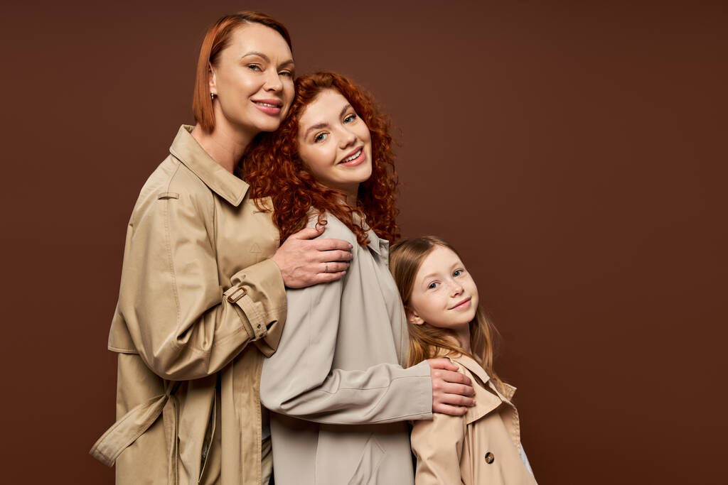 tre generazioni, donne rosse allegre e ragazza carina in trench posa su sfondo marrone - Foto, immagini