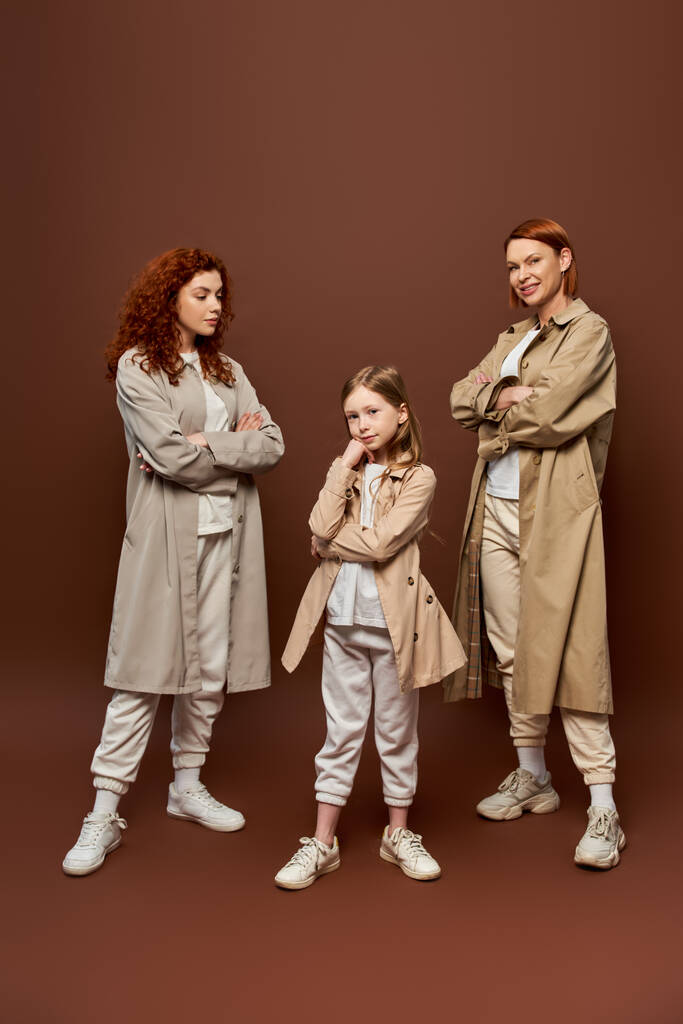 три покоління, жінки в пальто позують і дивляться на дівчину на коричневому тлі, руда сім'я - Фото, зображення