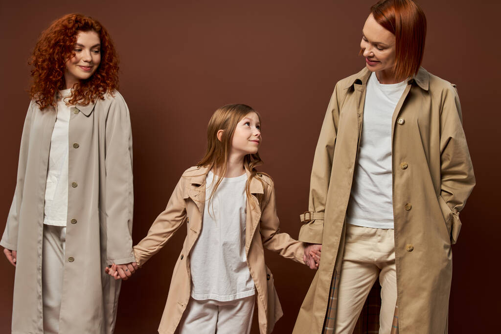 три покоління, весела руда сім'я в пальто тримає руки на коричневому тлі, жінки і дівчинка - Фото, зображення
