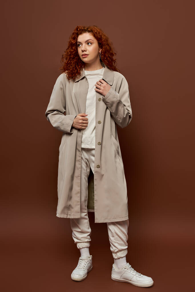 young woman with red curly hair posing in autumn coat and joggers on brown background, fall season - Photo, Image