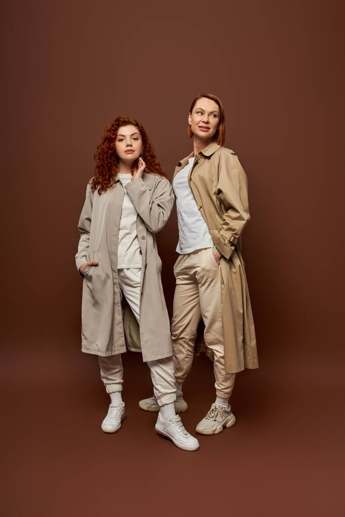 two generations, redhead women in trendy autumn clothes posing on brown background, trench coats - Photo, Image