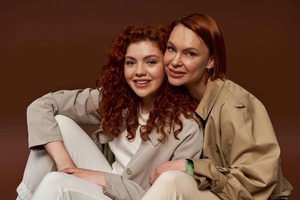 two generations, cheerful women with red hair posing in trendy autumn attire on brown background - Photo, Image