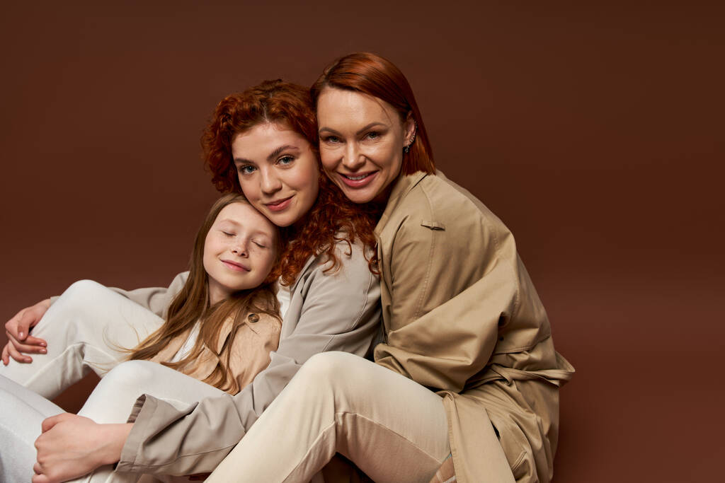 tres generaciones de mujeres, familia pelirroja positiva mirando a la cámara sobre fondo marrón, otoño - Foto, Imagen