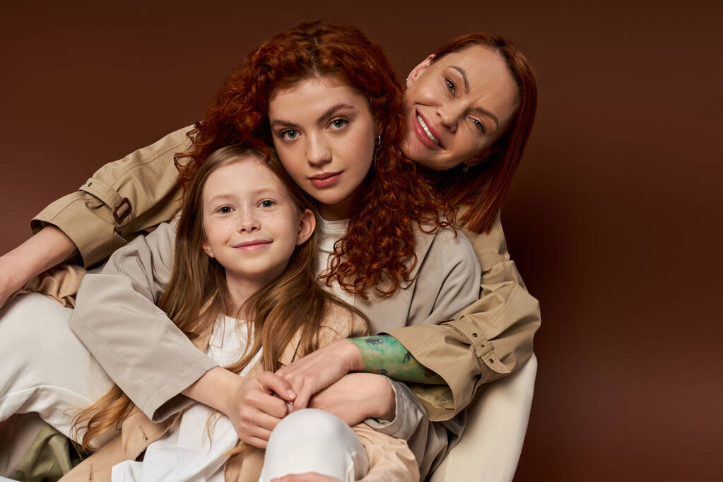 Üç nesil, pozitif kızıl saçlı aile kahverengi arka planda kameraya bakıyor, modern ebeveynlik. - Fotoğraf, Görsel