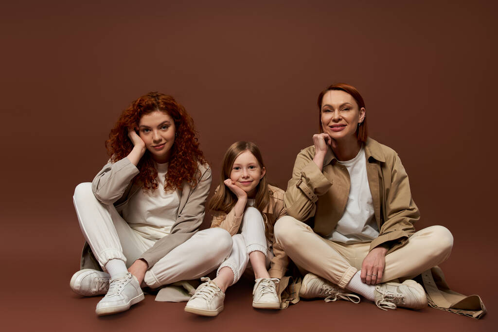 heureux rousse famille de trois générations féminines assis sur fond brun, manteaux d'automne - Photo, image
