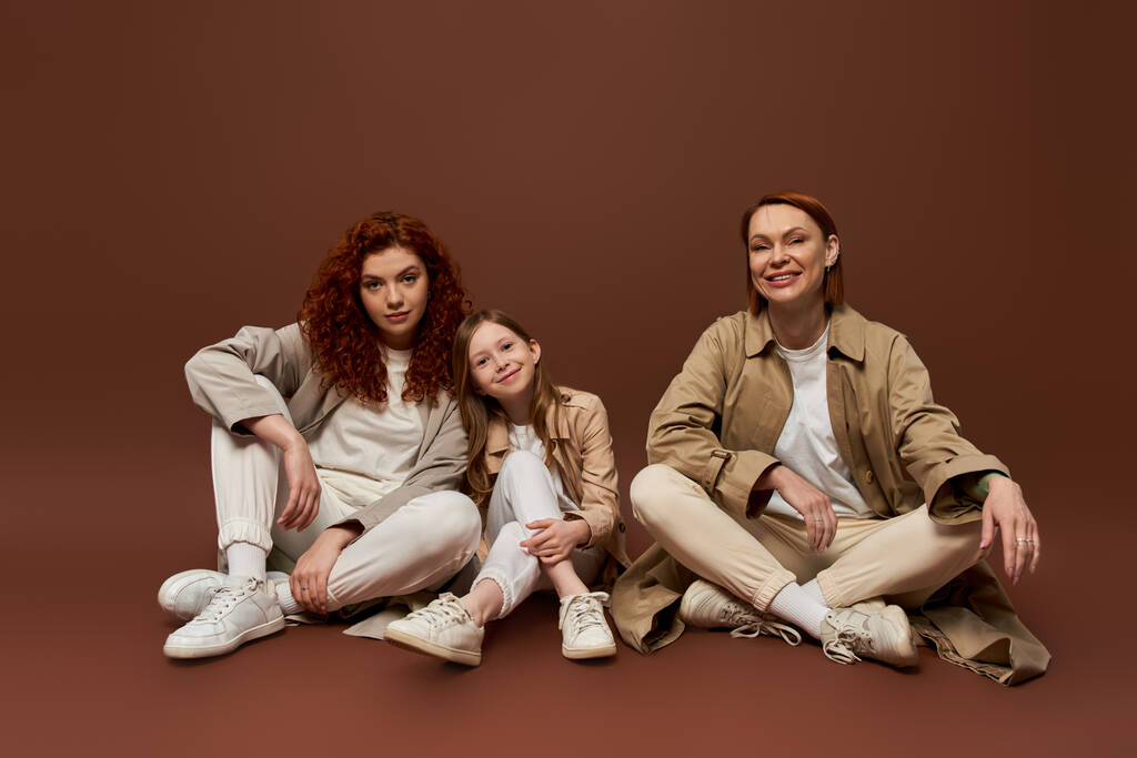 gioiosa famiglia rossa di tre generazioni femminili sedute su sfondo marrone, moda autunno - Foto, immagini