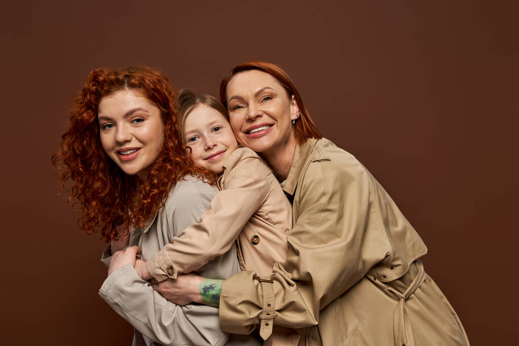 gelukkig roodharige familie van drie vrouwelijke generaties knuffelen op bruine achtergrond, herfst mode - Foto, afbeelding