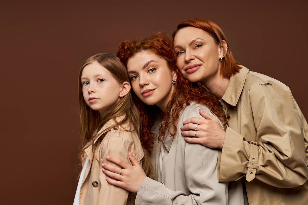 família de três gerações femininas com cabelos vermelhos posando em casacos bege, abraçando em fundo marrom - Foto, Imagem
