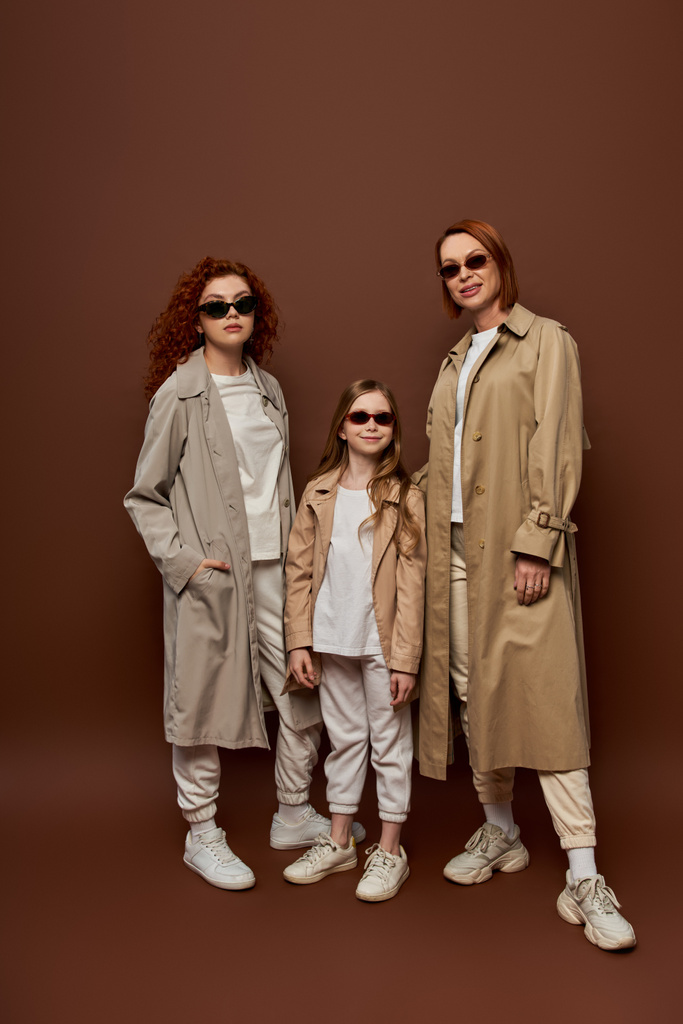 сімейна фотографія жіночих поколінь в сонцезахисних окулярах і пальто, позує на коричневому фоні, повна довжина - Фото, зображення