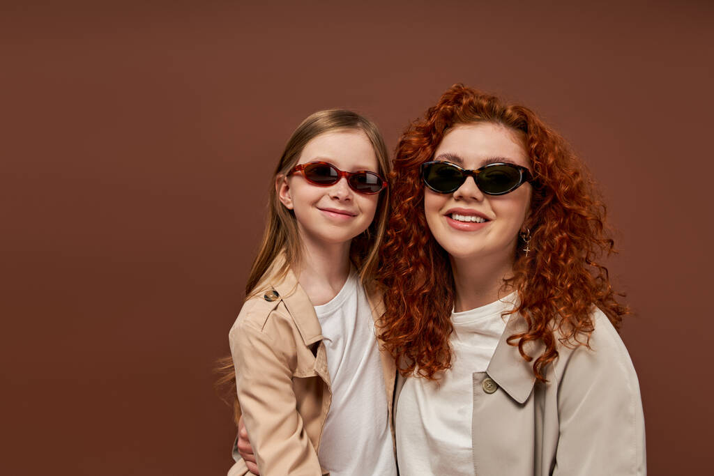 два покоління, щаслива кучерява мати і дочка в сонцезахисних окулярах на коричневому тлі, портрет - Фото, зображення
