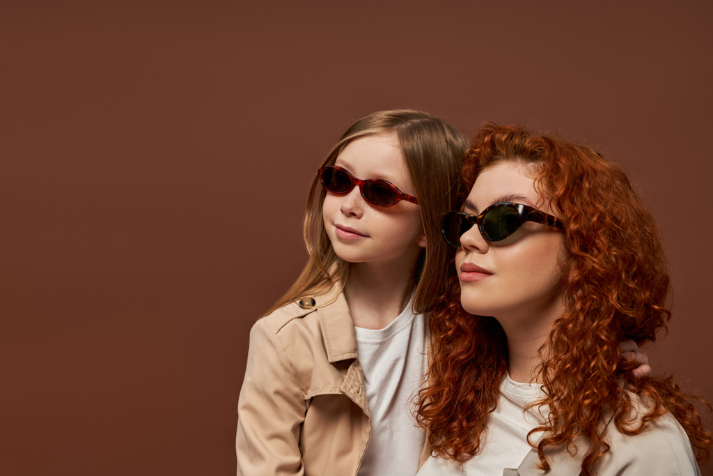 İki nesil, mutlu kızıl saçlı, güneş gözlüklü anne ve kız kahverengi arka plana bakıyorlar. - Fotoğraf, Görsel