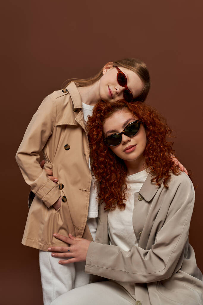 two generations, redhead mother and child in sunglasses and beige trench coats on brown backdrop - Photo, Image