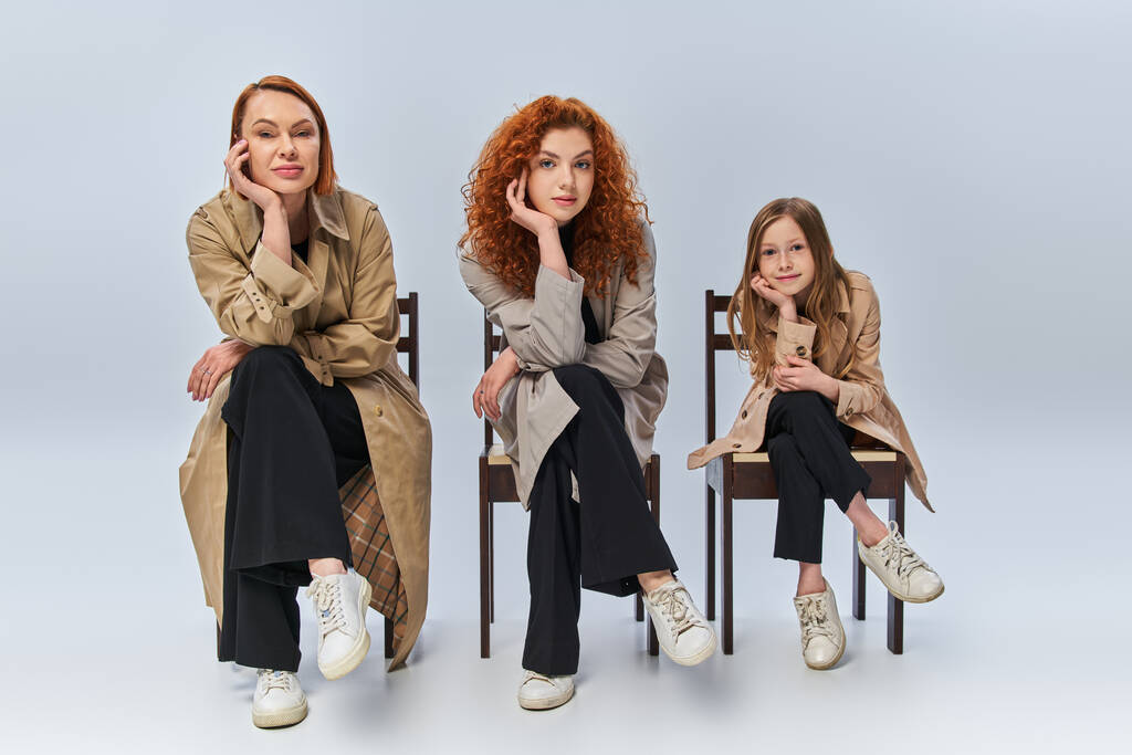 drei weibliche Generationen in Reihe auf Stühlen sitzend, glückliche rothaarige Frauen und Kind vor grauem Hintergrund - Foto, Bild