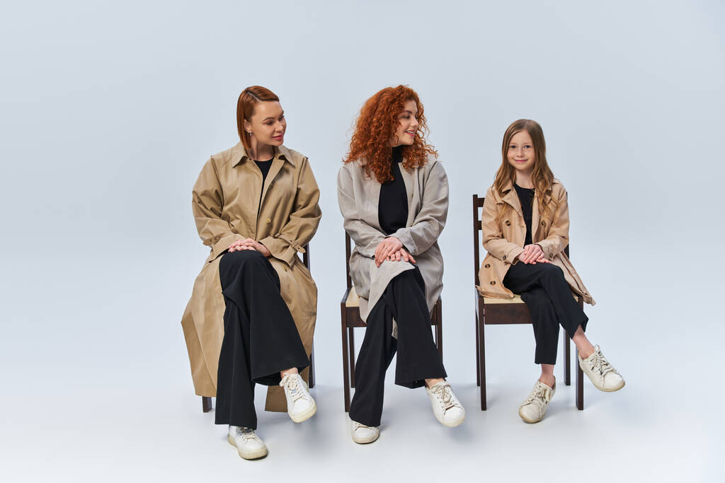 три покоління, руді жінки в пальто сидять на стільцях і дивляться на дівчину на сірому тлі - Фото, зображення