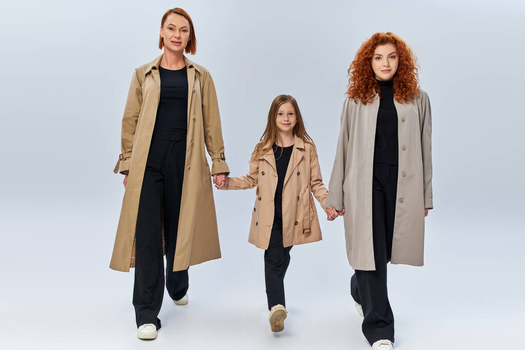 τρεις γυναίκες γενιές, κοκκινομάλλες γυναίκες και κορίτσι σε παλτά κρατώντας τα χέρια και περπατώντας σε γκρι φόντο - Φωτογραφία, εικόνα