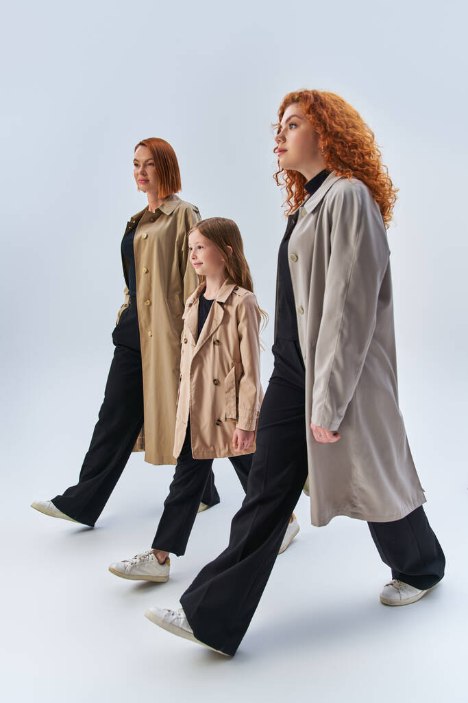 famille rousse de trois générations marchant avec les mains dans des poches de manteaux élégants sur fond gris - Photo, image