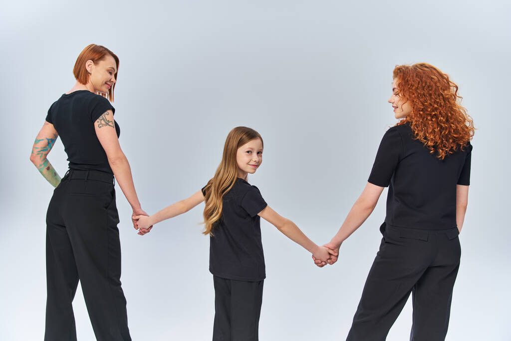 τρεις γενιές, ευτυχισμένη κοπέλα κρατώντας τα χέρια με τις γυναίκες, στέκεται στο ταίριασμα ενδυμασία σε γκρι φόντο - Φωτογραφία, εικόνα