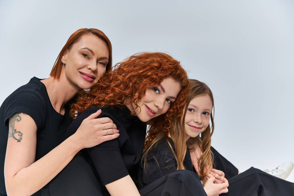 három generációs koncepció, vidám vörös hajú család mosolyog és szürke háttérrel nézi a kamerát - Fotó, kép