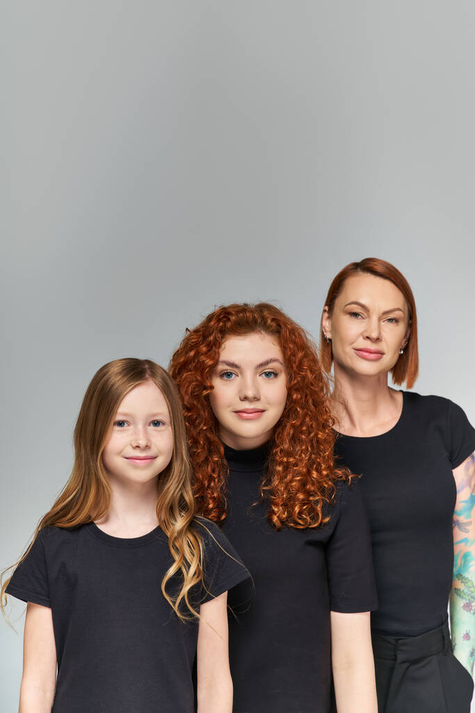 счастливые женщины рыжие волосы позируют с девушкой в одинаковых нарядах на сером фоне, три поколения - Фото, изображение