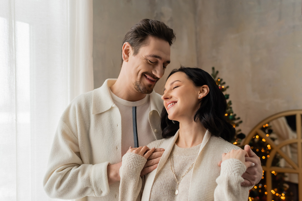 alegre hombre con barba abrazando feliz esposa en acogedor hogar desgaste cerca borrosa árbol de Navidad en el telón de fondo - Foto, Imagen
