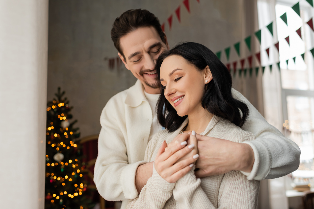 Keyifli bir adam sıcak bir evde karısına sarılıyor. Süslü bir Noel ağacının yanında, kış tatilinde. - Fotoğraf, Görsel
