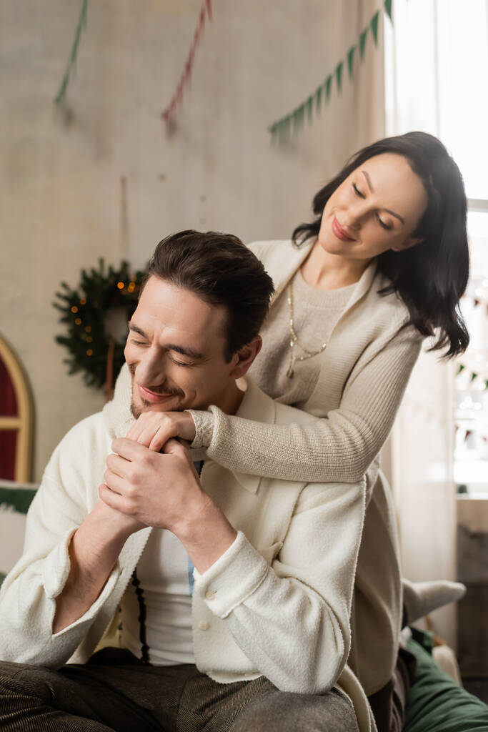 陽気な結婚したカップルは,現代アパートのクリスマスリース近くのベッドに座って手を握ります - 写真・画像
