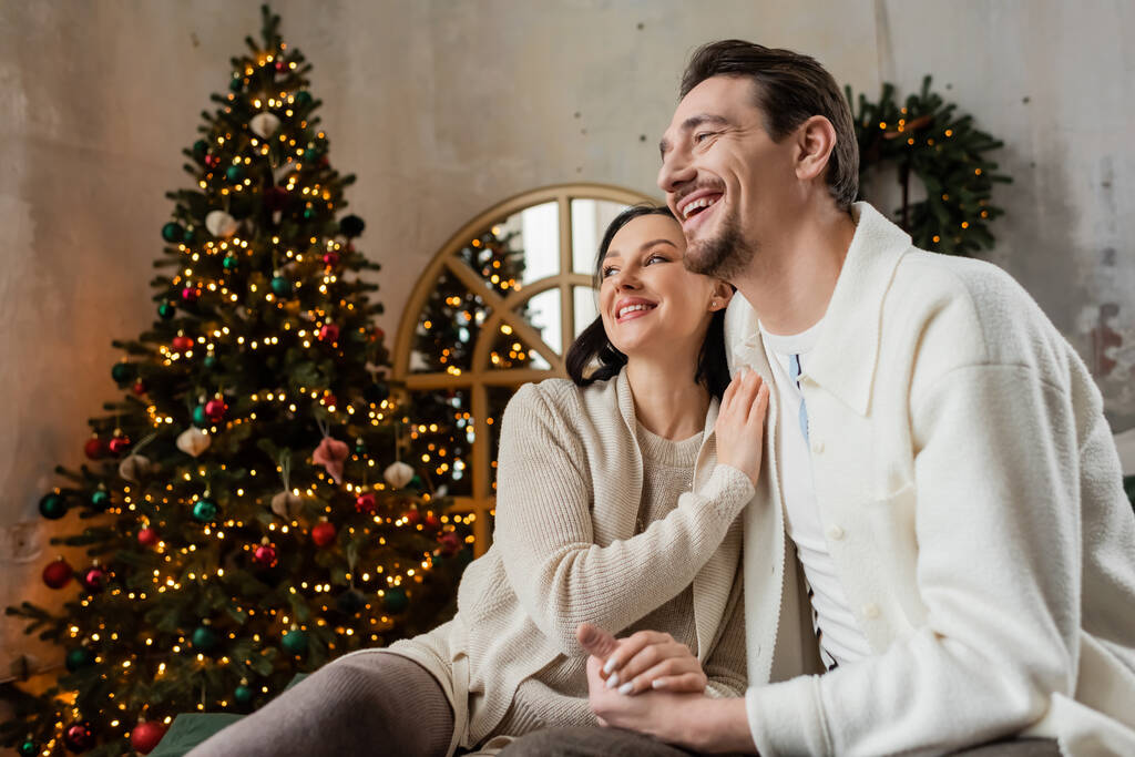 fröhlich verheiratetes Paar, Mann und Frau sitzen neben verschwommenem Weihnachtsbaum, Jahreszeit der Freude - Foto, Bild