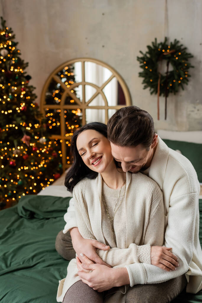夫は喜びの妻を受け入れて,ぼやけた装飾されたクリスマスツリーの近くのベッドに一緒に座っています - 写真・画像
