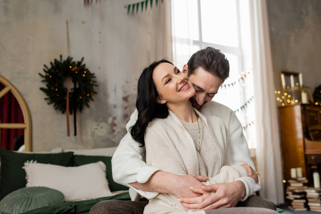 marido abrazando esposa alegre y sentados juntos en la cama cerca borrosa corona de Navidad en la pared - Foto, imagen