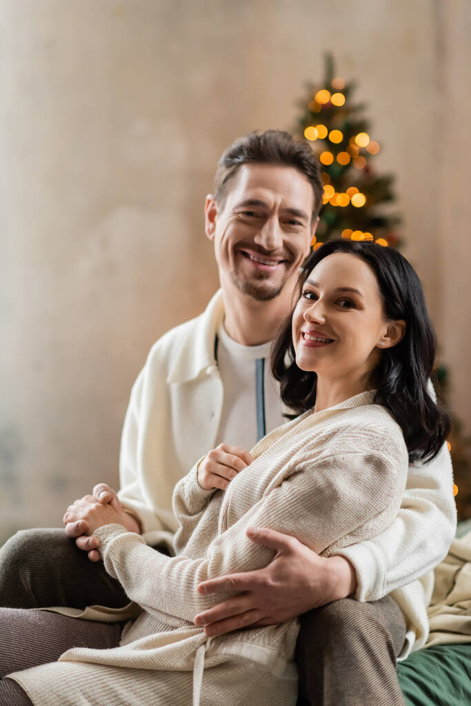cozy living, joyful married couple hugging each other near blurred Christmas lights on backdrop - Photo, Image
