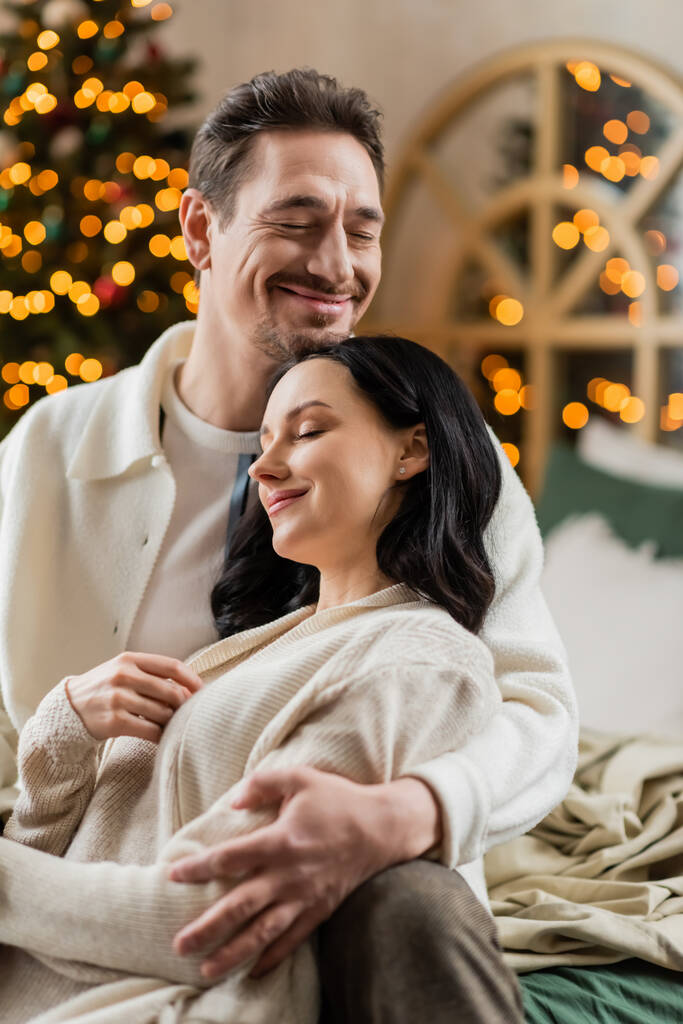 уютная жизнь, счастливая супружеская пара обнимающая друг друга возле размытых рождественских огней на заднем плане - Фото, изображение