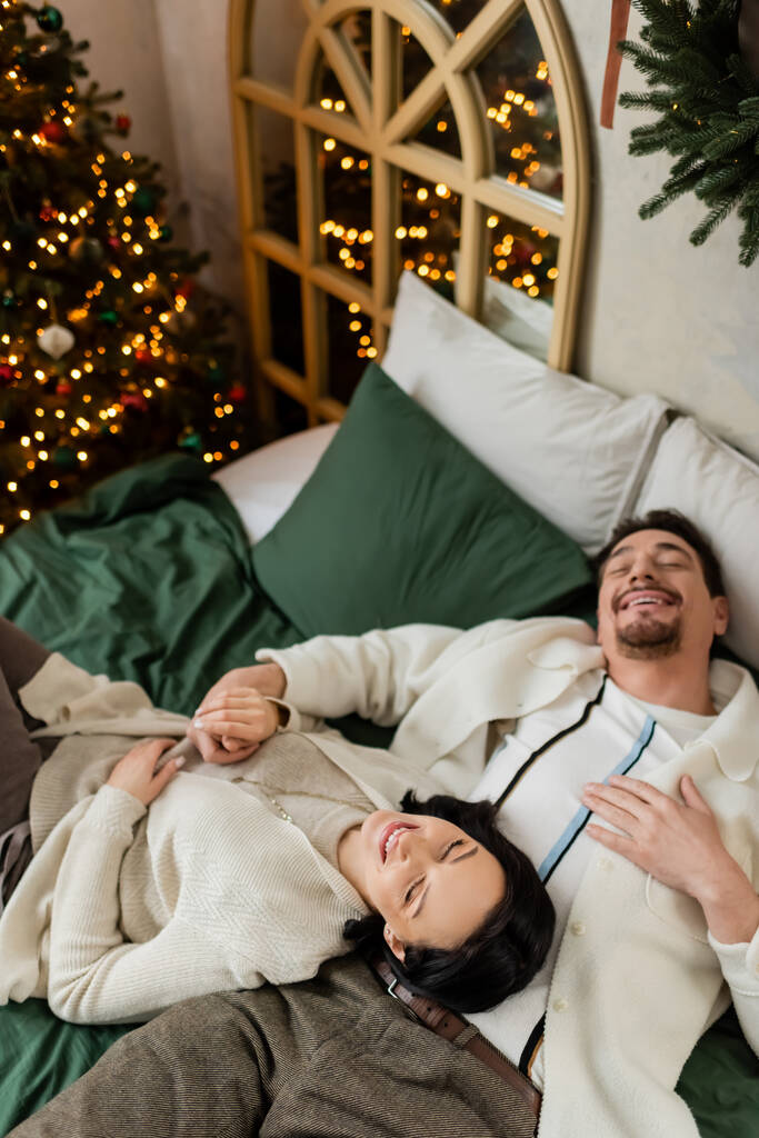 bovenaanzicht van vrolijk getrouwd stel brengt gezellige ochtend door in bed in de buurt van versierde kerstboom - Foto, afbeelding
