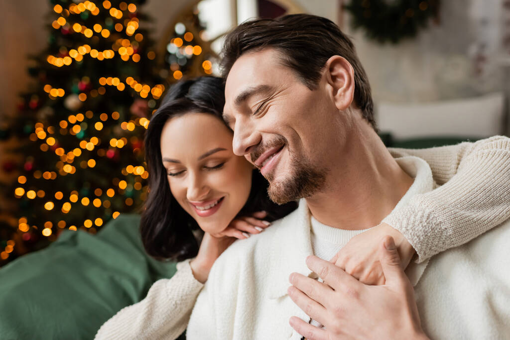 temporada de alegría, mujer feliz abrazando marido cerca del árbol de Navidad decorado en el dormitorio moderno - Foto, imagen