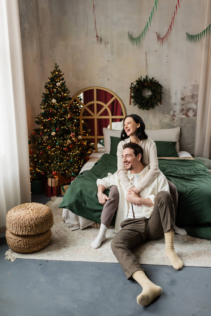 vacaciones de invierno, mujer feliz abrazando marido en dormitorio decorado con árbol de Navidad y corona - Foto, Imagen