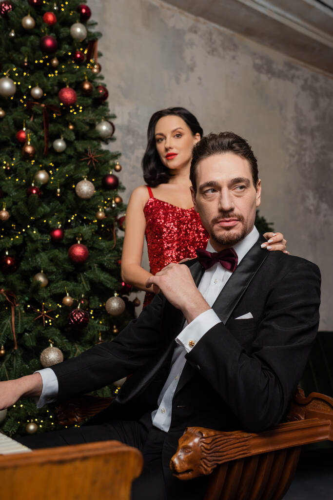 bohatý pár, hezká žena v červených šatech stojící poblíž manžela vedle zdobeného vánočního stromečku - Fotografie, Obrázek