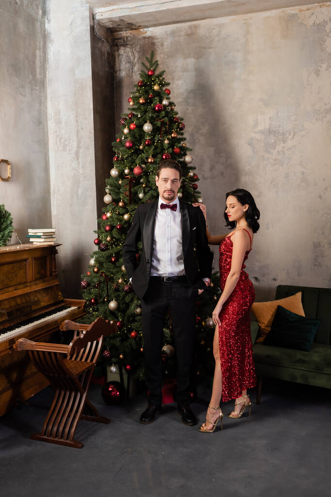 casal rico, mulher elegante em vestido vermelho de pé perto do homem em smoking, piano e árvore de Natal - Foto, Imagem