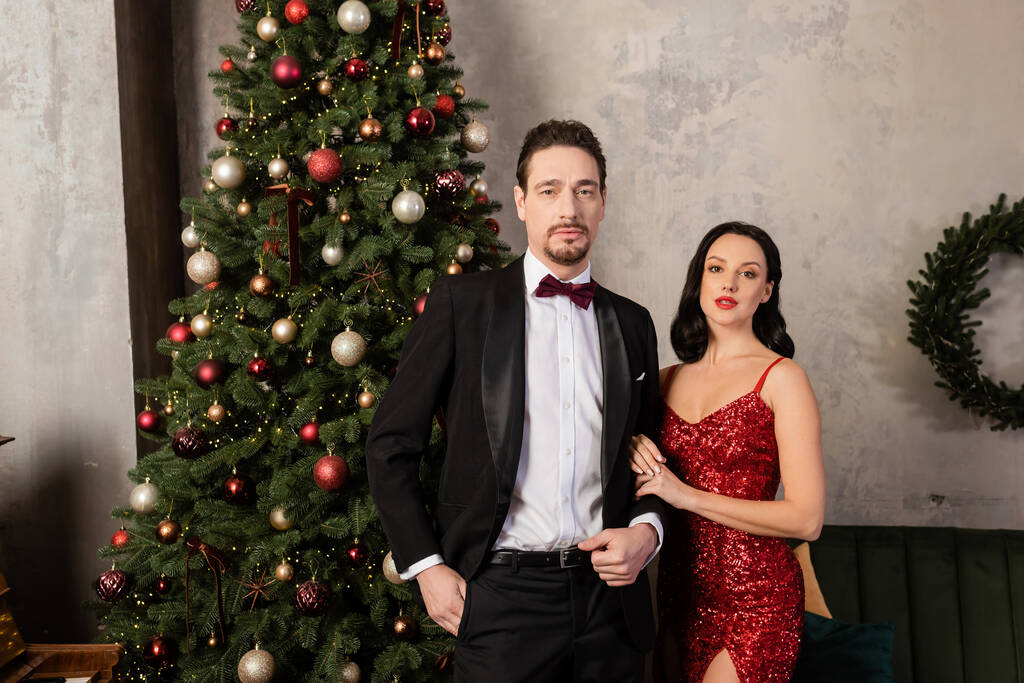 πλούσιο ζευγάρι, κομψή γυναίκα με κόκκινο φόρεμα στέκεται κοντά στον άνδρα με σμόκιν και διακοσμημένο χριστουγεννιάτικο δέντρο - Φωτογραφία, εικόνα