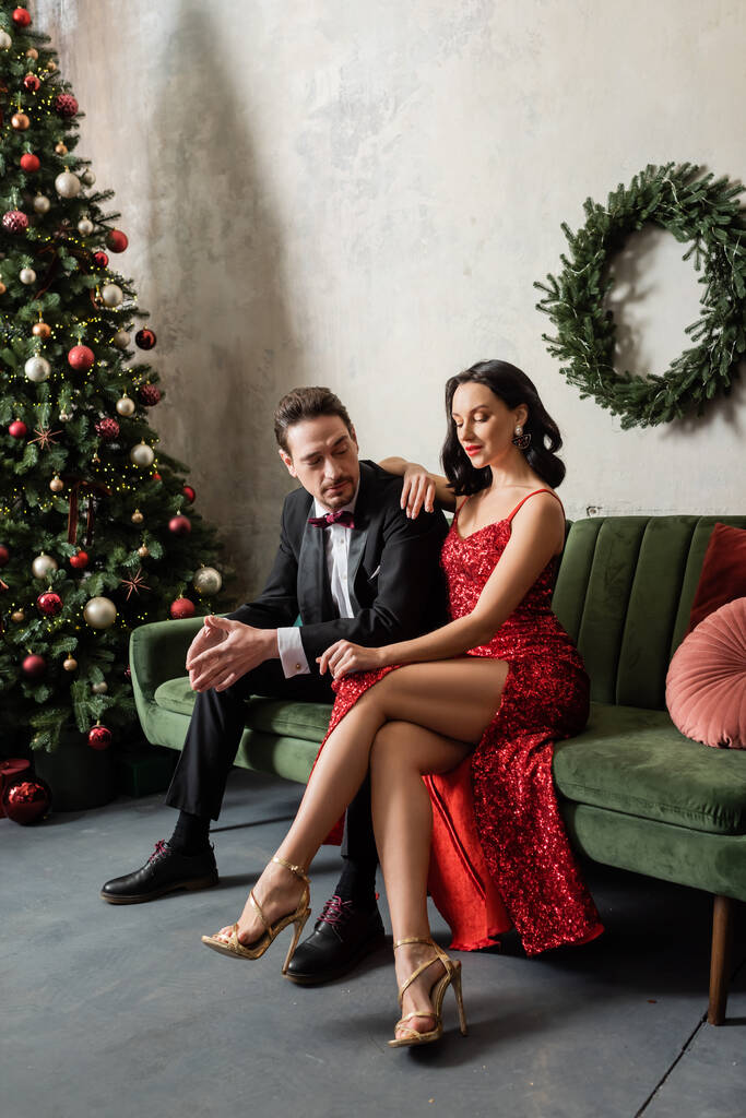 elegante y rica pareja en traje de noche sentado en el sofá cerca del árbol de Navidad decorado - Foto, imagen