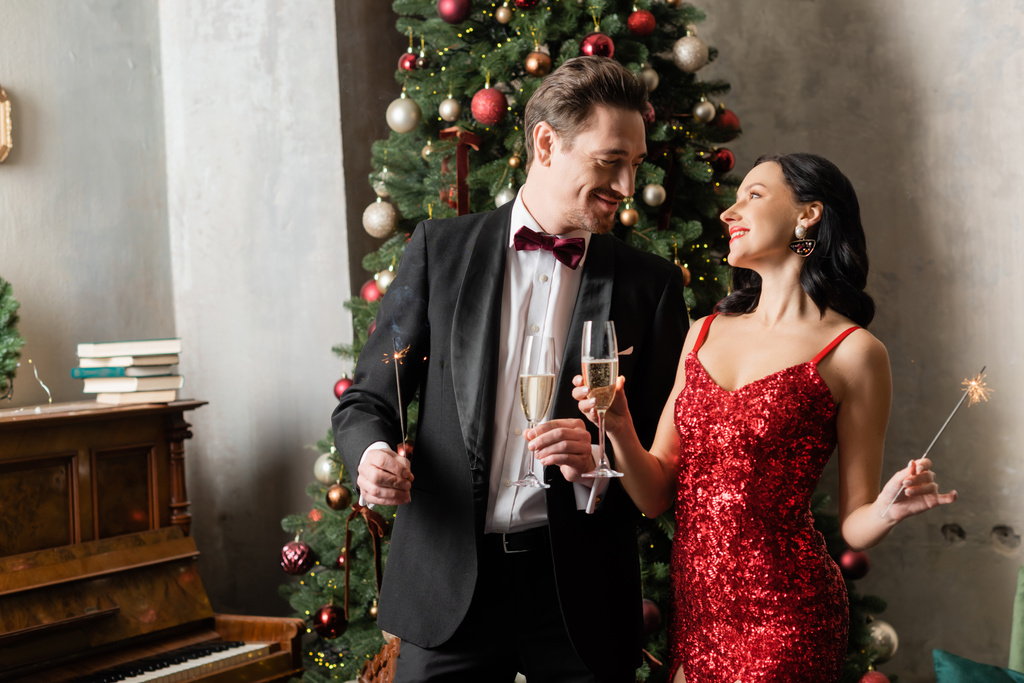 багата радісна пара в офіційному вбранні тримає келихи шампанського та блискавки біля ялинки - Фото, зображення
