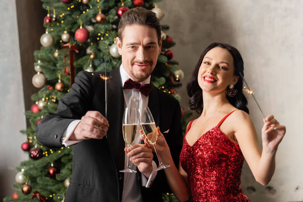 heureux couple marié en tenue formelle tenant des verres de champagne et des étincelles près de l'arbre de Noël - Photo, image