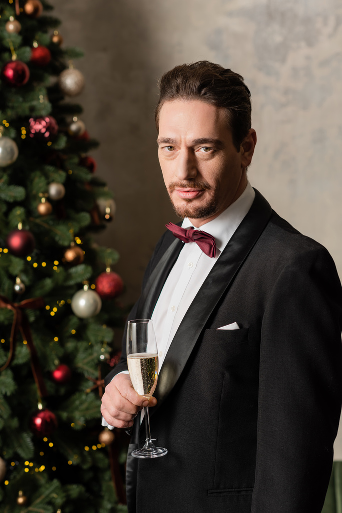 handsome wealthy gentleman in tuxedo holding champagne glass near decorated Christmas tree at home - Photo, Image