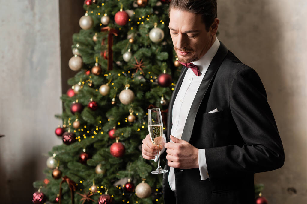 guapo hombre rico en esmoquin con pajarita sosteniendo copa de champán cerca del árbol de Navidad decorado - Foto, Imagen