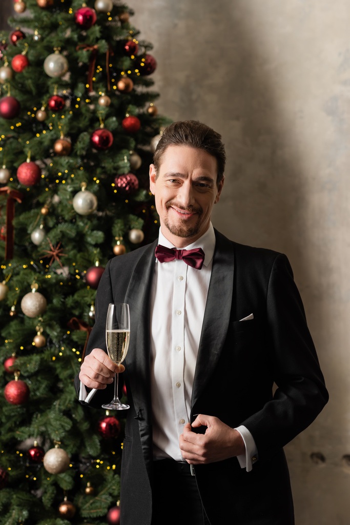 glücklicher reicher Mann im Smoking mit Fliege hält Champagnerglas neben geschmücktem Weihnachtsbaum - Foto, Bild