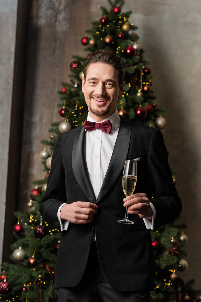 χαρούμενος κύριος με κοστούμι με παπιγιόν κρατώντας ποτήρι σαμπάνιας κοντά διακοσμημένο χριστουγεννιάτικο δέντρο - Φωτογραφία, εικόνα